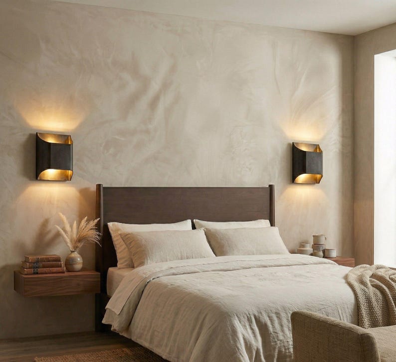 K&ouml;nnte beinhalten: Schlafzimmer-Interieur mit Bett, Nachttischen und Wandleuchten. Das Bett hat eine beige Bettdecke und Kissen. Zwei schwarze und goldene Wandleuchten sind an der Wand montiert. Ein Nachttisch aus Holz enth&auml;lt B&uuml;cher und eine Vase mit getrockneten Blumen.