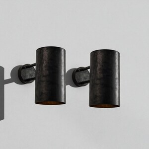 May include: Two dark metal wall sconces with cylindrical shades. Each sconce has a circular base and a curved arm connecting to the shade. The shades have a textured, aged appearance, and the interior glows with a warm light.