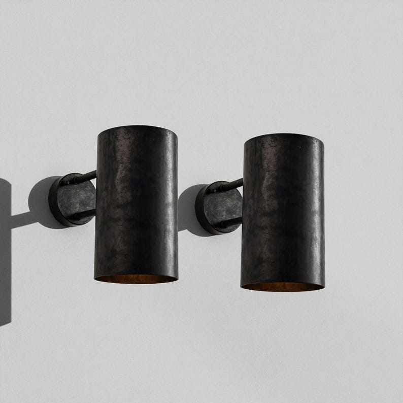 May include: Two dark metal wall sconces with cylindrical shades. Each sconce has a circular base and a curved arm connecting to the shade. The shades have a textured, aged appearance, and the interior glows with a warm light.
