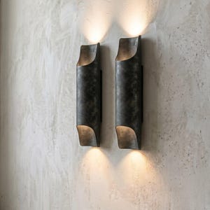 May include: Two dark metal wall sconces with a cylindrical design and a curved top and bottom. The sconces are mounted on a textured, light-colored wall, and each emits a warm, downward-facing light.