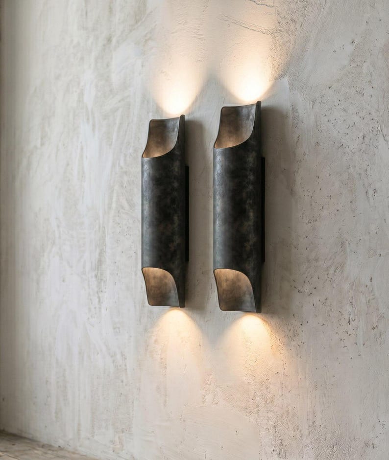 May include: Two dark metal wall sconces with a cylindrical design and a curved top and bottom. The sconces are mounted on a textured, light-colored wall, and each emits a warm, downward-facing light.