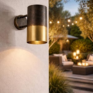 May include: A cylindrical outdoor wall sconce with a two-tone finish. The top portion is dark bronze, and the bottom is gold. The sconce is mounted on a textured white wall, with a warm light illuminating the surface below. The background shows an outdoor patio setting.