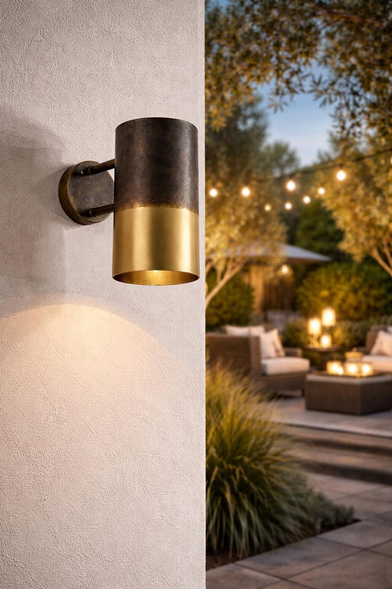 May include: A cylindrical outdoor wall sconce with a two-tone finish. The top portion is dark bronze, and the bottom is gold. The sconce is mounted on a textured white wall, with a warm light illuminating the surface below. The background shows an outdoor patio setting.