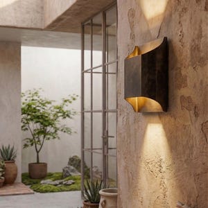 K&ouml;nnte beinhalten: Eine bronzefarbene Au&szlig;enwandleuchte mit geschwungenem Design und goldenem Innenraum. Die Leuchte ist an einer strukturierten, hellbraunen Wand angebracht und wirft einen Lichtstrahl nach unten. Die Szene umfasst Topfpflanzen und einen Blick auf eine Au&szlig;enterrasse.