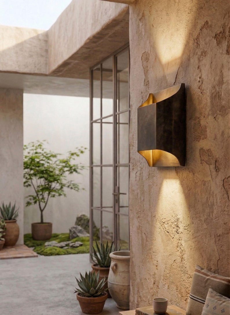 K&ouml;nnte beinhalten: Eine bronzefarbene Au&szlig;enwandleuchte mit geschwungenem Design und goldenem Innenraum. Die Leuchte ist an einer strukturierten, hellbraunen Wand angebracht und wirft einen Lichtstrahl nach unten. Die Szene umfasst Topfpflanzen und einen Blick auf eine Au&szlig;enterrasse.