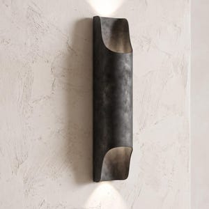May include: A dark, cylindrical wall sconce with a curved top and bottom, mounted on a textured white wall. The sconce emits a warm light from both ends, creating an inviting ambiance. The fixture has a metallic, weathered finish.