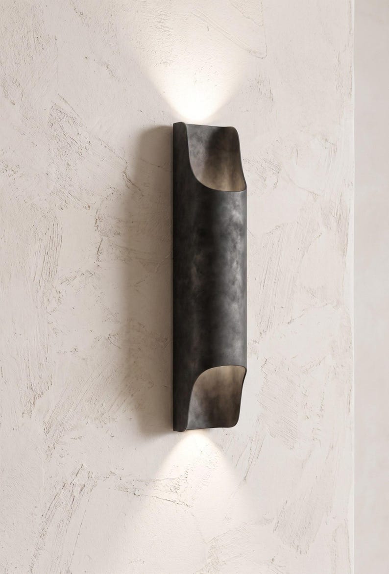 May include: A dark, cylindrical wall sconce with a curved top and bottom, mounted on a textured white wall. The sconce emits a warm light from both ends, creating an inviting ambiance. The fixture has a metallic, weathered finish.