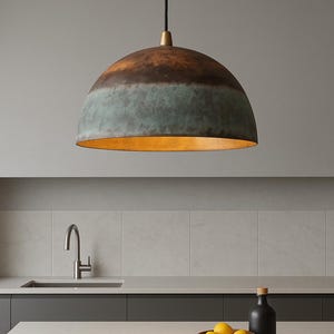 May include: A dome-shaped pendant light with a copper and teal finish hangs above a kitchen countertop. The interior of the light fixture has a warm, golden hue. The kitchen features a stainless steel faucet, a wooden bowl of lemons, and a black bottle.