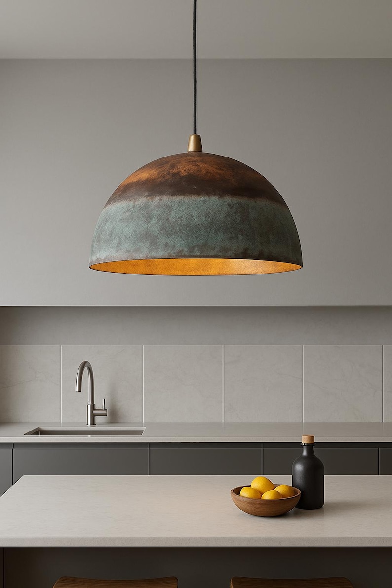 May include: A dome-shaped pendant light with a copper and teal finish hangs above a kitchen countertop. The interior of the light fixture has a warm, golden hue. The kitchen features a stainless steel faucet, a wooden bowl of lemons, and a black bottle.