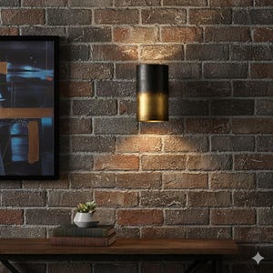 May include: A cylindrical wall sconce with a black top and a gold-colored bottom is mounted on a brick wall. The sconce is illuminated, casting a warm glow on the wall. The sconce is a decorative lighting fixture.