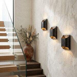 K&ouml;nnte beinhalten: Drei dunkelbronze Wandleuchten mit warmem, goldenem Schein sind an einer strukturierten, cremefarbenen Wand angebracht. Eine gro&szlig;e, rustikale Terrakotta-Vase mit getrockneten Pflanzen steht auf einer Holztreppe.