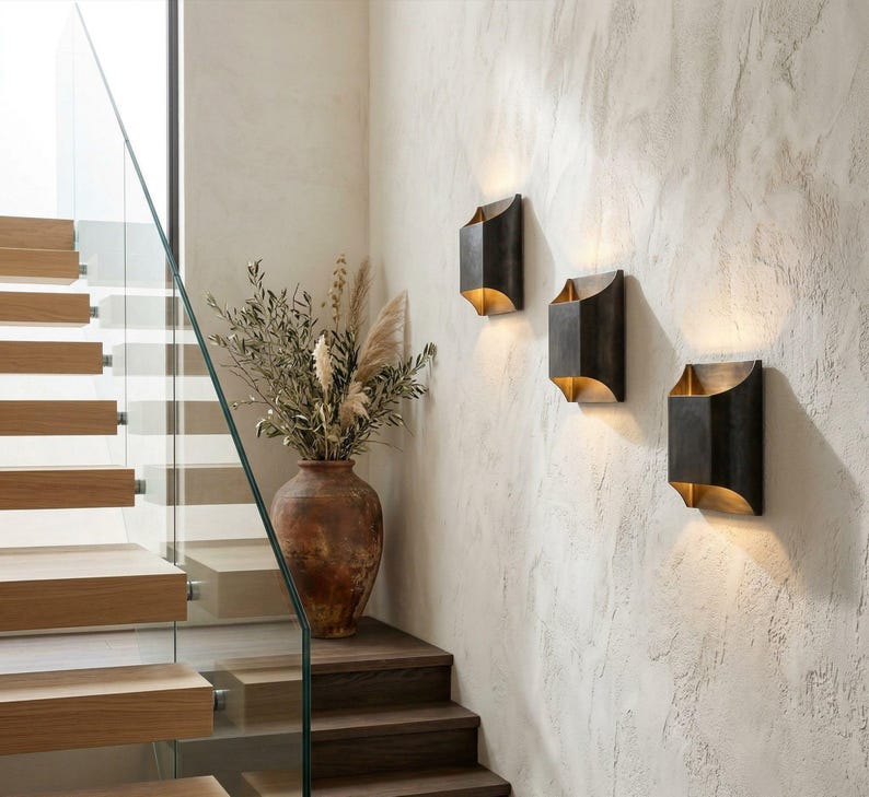 K&ouml;nnte beinhalten: Drei dunkelbronze Wandleuchten mit warmem, goldenem Schein sind an einer strukturierten, cremefarbenen Wand angebracht. Eine gro&szlig;e, rustikale Terrakotta-Vase mit getrockneten Pflanzen steht auf einer Holztreppe.