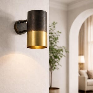 May include: A cylindrical wall sconce with a two-tone finish. The top portion is dark bronze, and the bottom is gold. The sconce is mounted on a circular bronze base. The light shines downward, illuminating the wall below.
