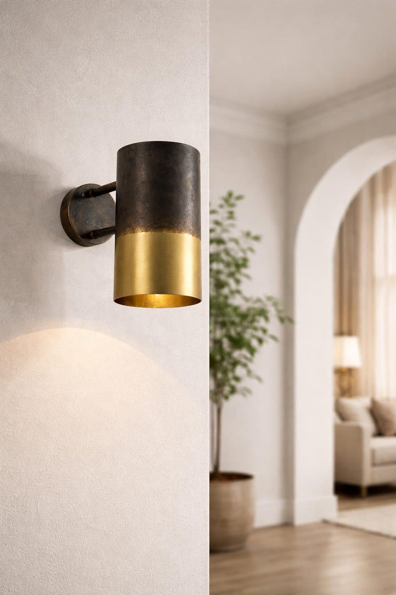 May include: A cylindrical wall sconce with a two-tone finish. The top portion is dark bronze, and the bottom is gold. The sconce is mounted on a circular bronze base. The light shines downward, illuminating the wall below.