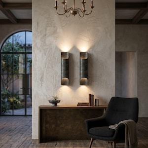 May include: Two dark metal wall sconces with a cylindrical design are mounted on a textured wall. A small bronze chandelier hangs above. A wooden console table holds a decorative bowl and books. A black armchair with a blanket sits nearby.