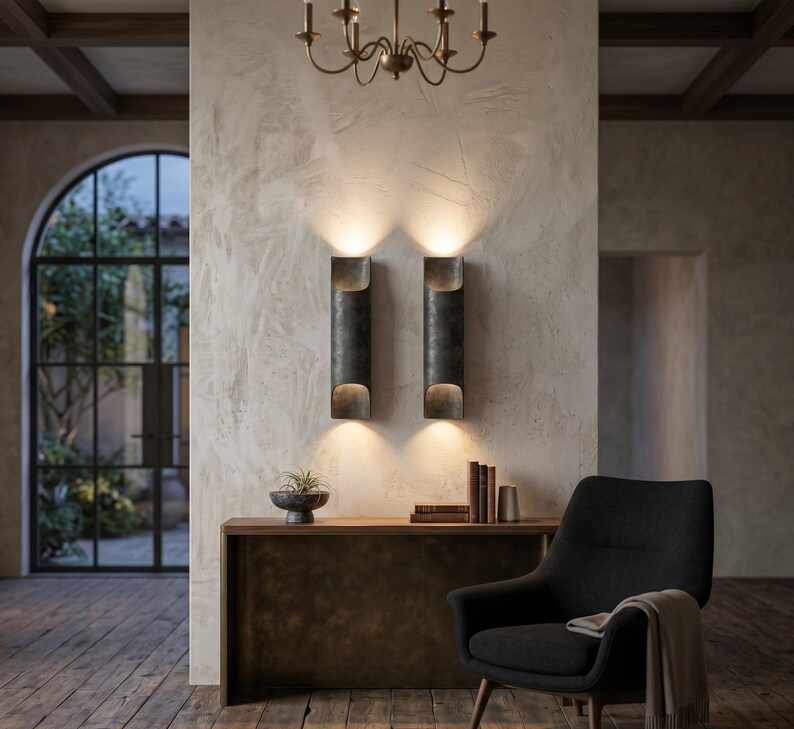 May include: Two dark metal wall sconces with a cylindrical design are mounted on a textured wall. A small bronze chandelier hangs above. A wooden console table holds a decorative bowl and books. A black armchair with a blanket sits nearby.