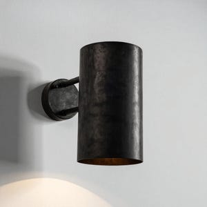 May include: A cylindrical wall sconce with a dark, textured finish. The sconce has a circular base and a curved arm that connects to the cylindrical shade. The interior of the shade has a warm, golden hue. The fixture is mounted on a white wall.