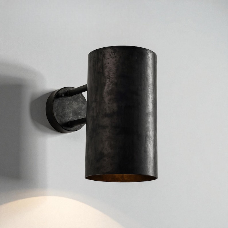 May include: A cylindrical wall sconce with a dark, textured finish. The sconce has a circular base and a curved arm that connects to the cylindrical shade. The interior of the shade has a warm, golden hue. The fixture is mounted on a white wall.