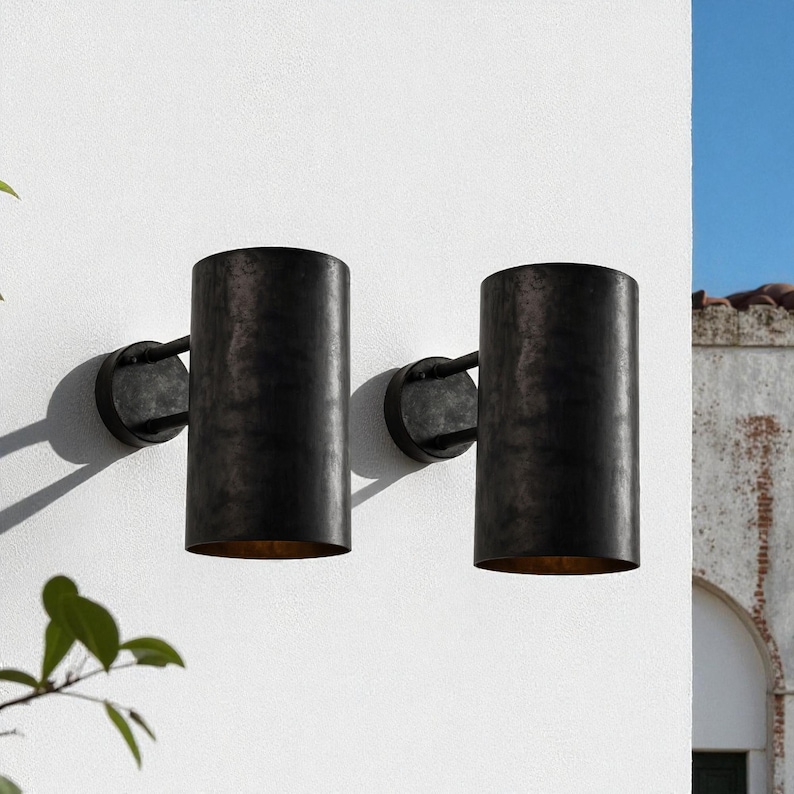 May include: Two cylindrical, dark bronze outdoor wall sconces mounted on a white wall. Each sconce has a circular base and a long, vertical shade. The sconces are made of metal and have a weathered finish, suggesting an aged appearance.