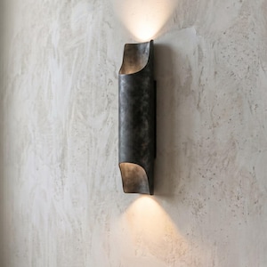 May include: A dark gray, cylindrical wall sconce with a curved top and bottom. The sconce emits a warm light upwards and downwards, creating a soft glow. The fixture is mounted on a textured, light gray wall.