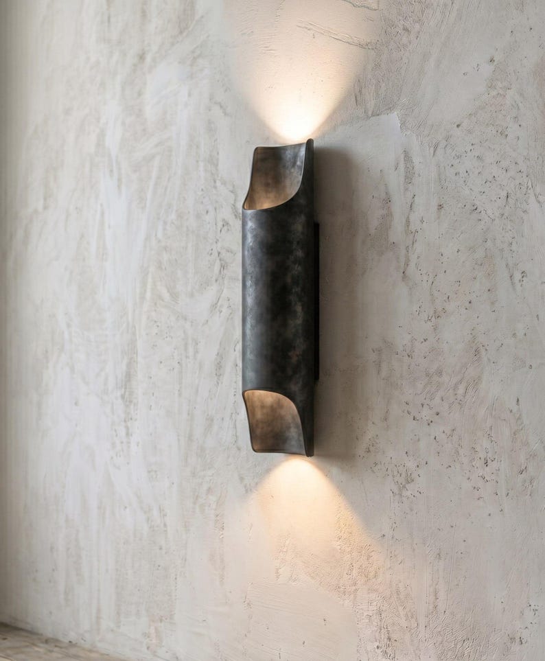 May include: A dark gray, cylindrical wall sconce with a curved top and bottom. The sconce emits a warm light upwards and downwards, creating a soft glow. The fixture is mounted on a textured, light gray wall.