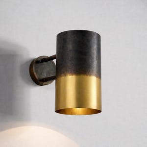 May include: A cylindrical wall sconce with a two-tone finish. The top half is dark gray, transitioning to a gold-colored bottom. The sconce is mounted on a circular base with two supporting arms.