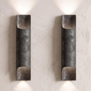 May include: Two dark gray, cylindrical wall sconces with a textured finish are mounted on a textured white wall. Each sconce has an open top and bottom, emitting a warm light. The sconces are evenly spaced.