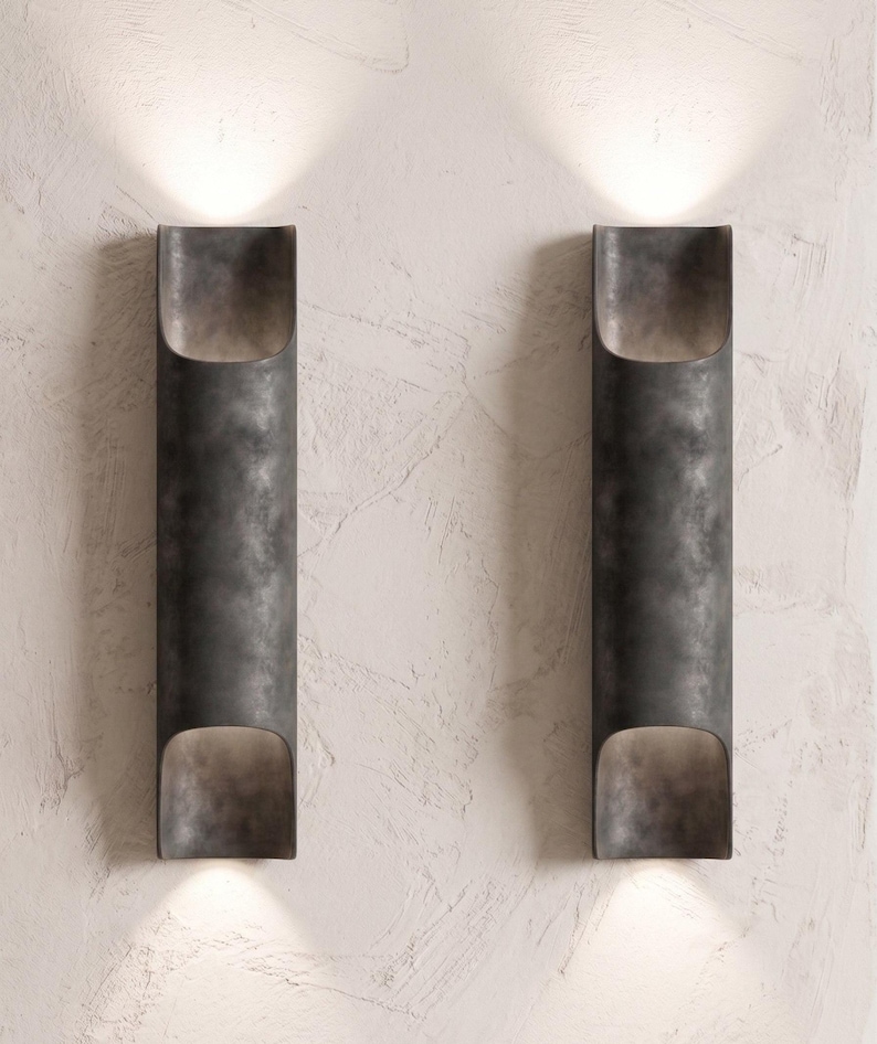 May include: Two dark gray, cylindrical wall sconces with a textured finish are mounted on a textured white wall. Each sconce has an open top and bottom, emitting a warm light. The sconces are evenly spaced.