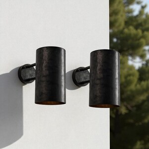 May include: Two cylindrical, dark bronze outdoor wall sconces. Each sconce has a circular base and a cylindrical shade. The sconces are mounted on a white wall, with a blurred background of green foliage.