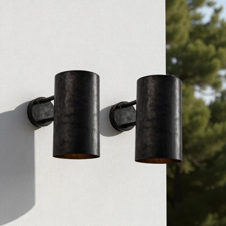 May include: Two cylindrical, dark bronze outdoor wall sconces. Each sconce has a circular base and a cylindrical shade. The sconces are mounted on a white wall, with a blurred background of green foliage.
