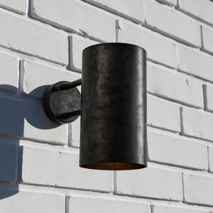 May include: A cylindrical, dark metal outdoor wall sconce. The sconce is mounted on a circular base with a curved arm. The fixture is attached to a white brick wall. The interior of the sconce is a warm, golden color.