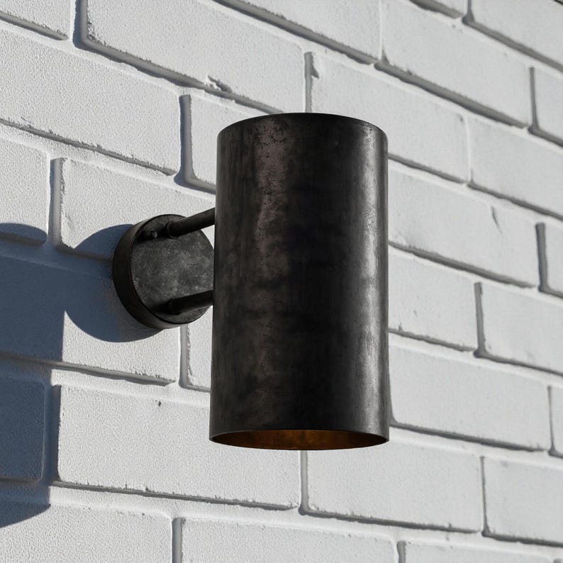 May include: A cylindrical, dark metal outdoor wall sconce. The sconce is mounted on a circular base with a curved arm. The fixture is attached to a white brick wall. The interior of the sconce is a warm, golden color.