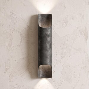 May include: A dark gray, cylindrical wall sconce with a textured finish. The sconce has an open top and bottom, emitting light upwards and downwards. The fixture is mounted on a textured, off-white wall.