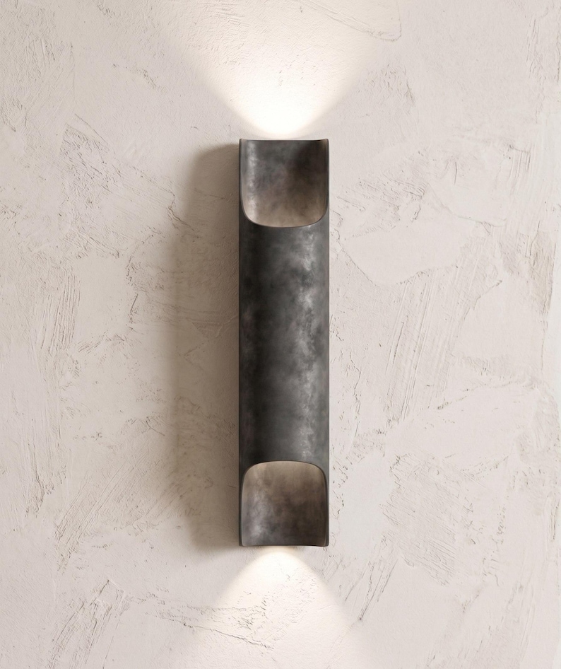 May include: A dark gray, cylindrical wall sconce with a textured finish. The sconce has an open top and bottom, emitting light upwards and downwards. The fixture is mounted on a textured, off-white wall.