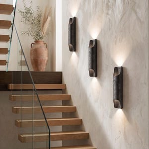 May include: Three dark metal wall sconces with a warm glow are mounted on a textured, light-colored wall. A wooden staircase with glass railing and a terracotta vase with dried plants are also visible.