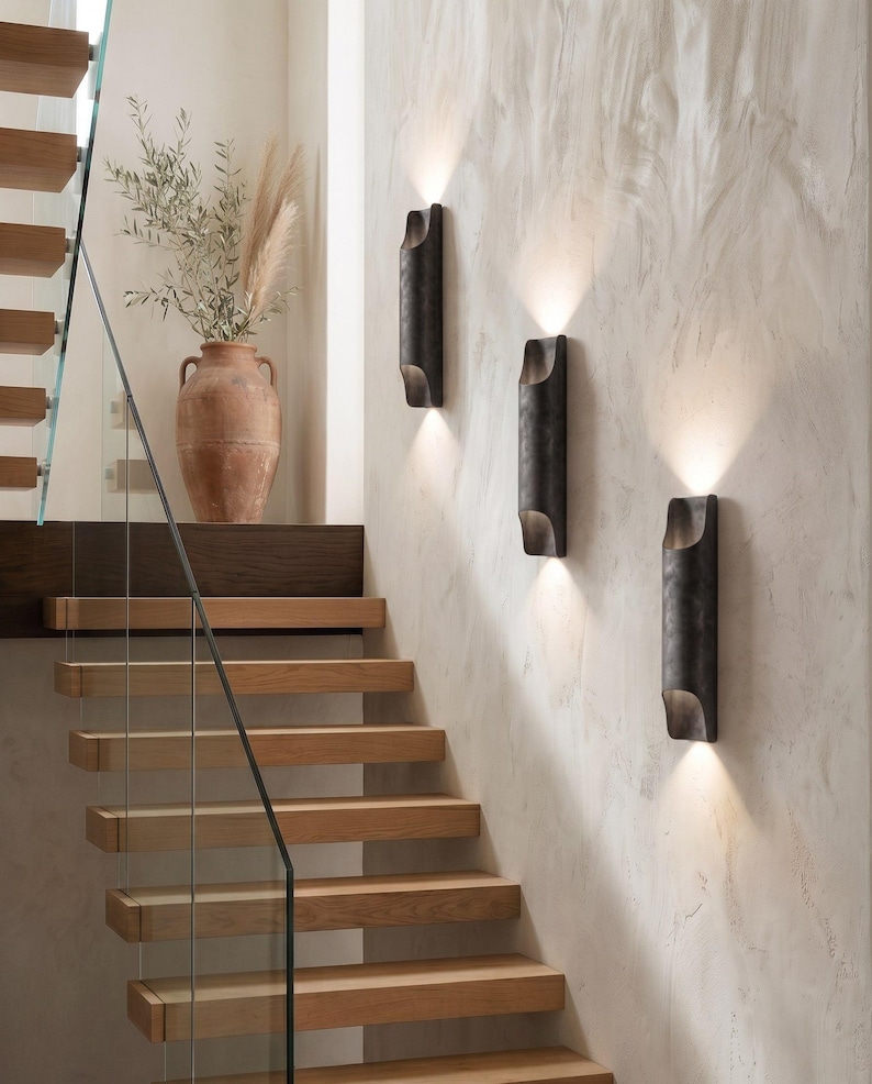 May include: Three dark metal wall sconces with a warm glow are mounted on a textured, light-colored wall. A wooden staircase with glass railing and a terracotta vase with dried plants are also visible.