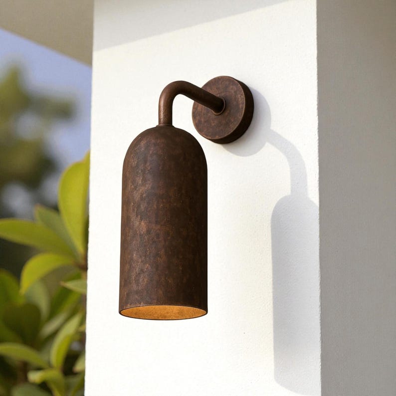 Pu&ograve; includere: Applique da parete per esterni in bronzo. Il dispositivo presenta un paralume cilindrico strutturato e un braccio curvo che lo collega a una piastra di montaggio circolare. La luce &egrave; montata su un muro bianco, proiettando un'ombra.