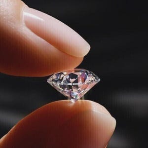 May include: A sparkling, brilliant-cut diamond held between two fingers. The diamond's facets reflect light, creating a dazzling effect. The background is a dark colour, highlighting the diamond's clarity. The image focuses on the diamond.