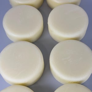 Pode incluir: Oito sabonetes redondos, esbranquiçados, dispostos em duas filas sobre uma superfície azul clara. As barras têm um aspeto liso e ligeiramente texturizado, sugerindo um produto natural ou feito à mão. A iluminação realça a forma e a textura do sabonete.