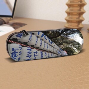 May include: A black zippered pencil case with a Japanese-themed design. The case features a print of white paper lanterns with blue Japanese characters. The background of the case shows a tree-lined path.