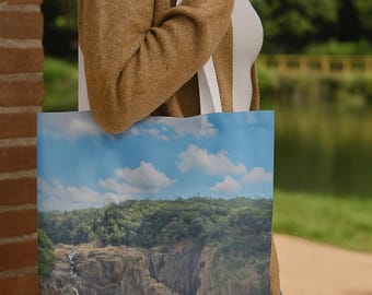 May include: A tote bag featuring a scenic landscape of a waterfall cascading down a rocky cliff, under a blue sky with fluffy white clouds. The bag has white straps and is being carried.