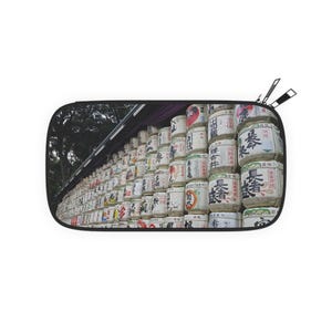 May include: A rectangular travel pouch featuring a photograph of a wall of sake barrels. The barrels are white with black and red Japanese characters. The pouch has a black border and two zippers.
