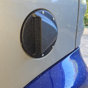 Puede incluir: Primer plano de la tapa del depósito de combustible de un coche. La puerta negra y redonda tiene una superficie texturizada y un diseño triangular. Está fijada a una carrocería plateada y azul con pequeños tornillos.