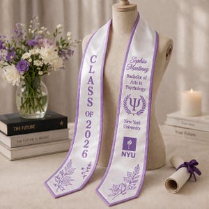 May include: A white graduation sash with lilac trim and text. The sash reads "CLASS OF 2026" and includes the name "Sophia Martinez", "Bachelor of Arts in Psychology", and "New York University". Decorative floral elements are also present.
