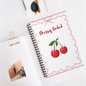 May include: A spiral-bound notebook with a white cover featuring a red scalloped border and the words "Cherry Coded" in red script. The cover also displays a pair of red cherries with green leaves and pink bows. Sunglasses and a gold binder clip are also visible.