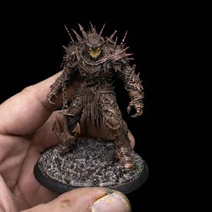 May include: A detailed miniature figure of a menacing, armored creature with spikes and a chain weapon. The figure is painted in shades of brown and bronze, with a menacing yellow-green glow emanating from its face. The figure is mounted on a textured, gray base.