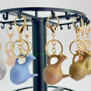 May include: A selection of miniature watering can keyrings in shades of blue, brown, and gold. Each keyring is attached to a gold-coloured clasp and ring, suspended from a black display stand.