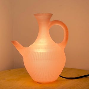 May include: A pink, glowing jug-shaped lamp with a handle and spout. The lamp has vertical ribbed detailing and is illuminated from within, casting a warm, soft light. The lamp is on a wooden surface.