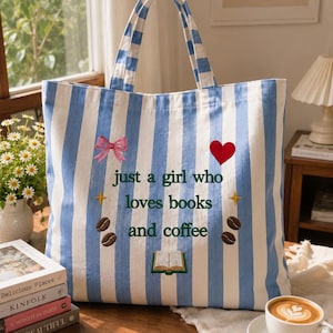 Op de afbeelding: Een blauw-wit gestreepte tote bag met geborduurde ontwerpen, waaronder een roze strik, een rood hart, twee gele sterren, vier koffiebonen en een open boek. De tekst "just a girl who loves books and coffee" is in het donkergroen geborduurd.