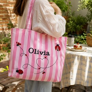 Könnte beinhalten: Eine rosa-weiß gestreifte Tragetasche mit gestickten roten Marienkäfern und einer gepunkteten Linie, die ein Herz bildet. Der Name Olivia ist in schwarzer Schrift in der Mitte der Tasche eingestickt. Die Tasche hat zwei lange Trageriemen.